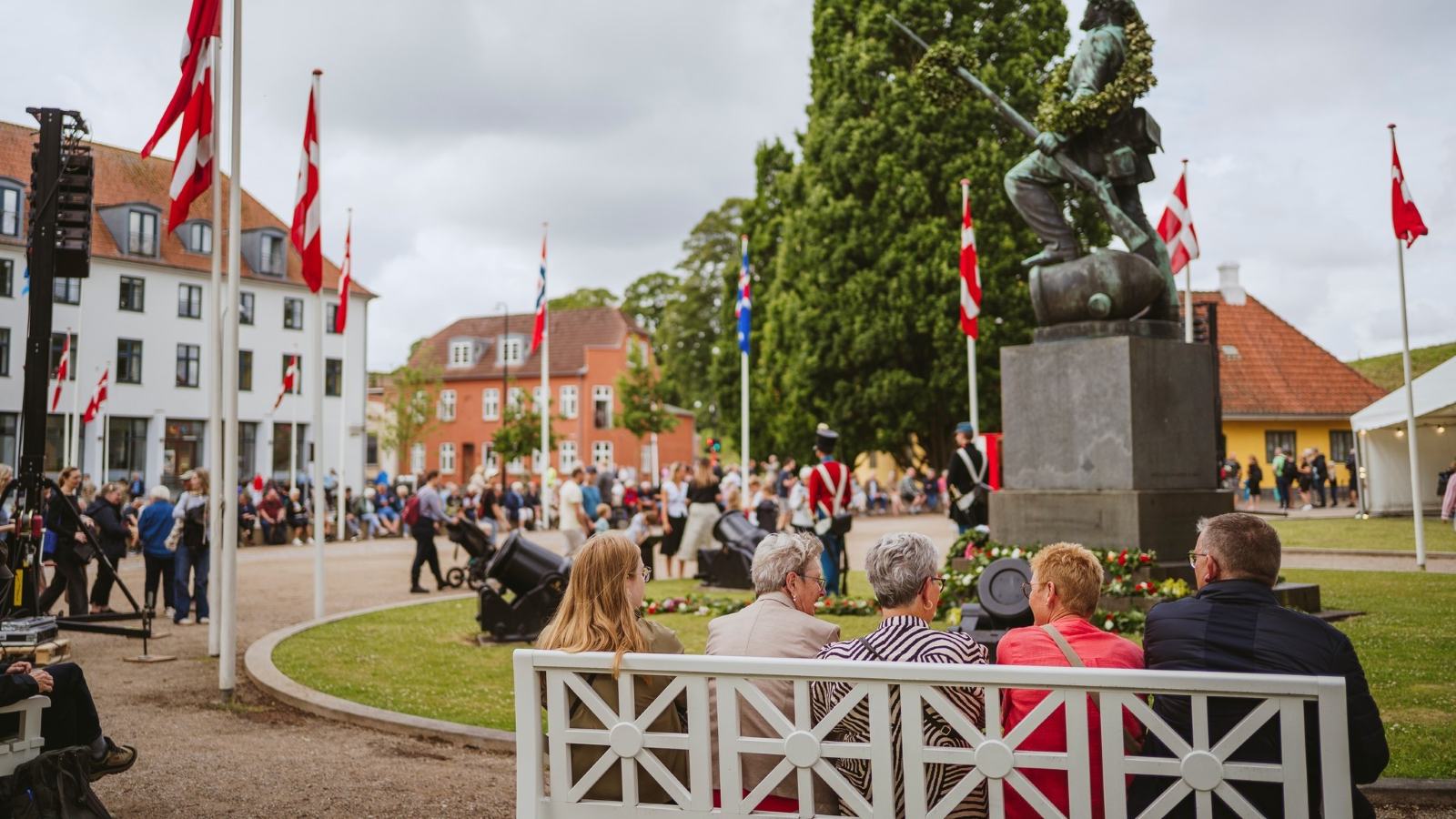 Mennesker sidder på bænk på Landsoldatpladsen og kigger på optog til Fæstningsdagene